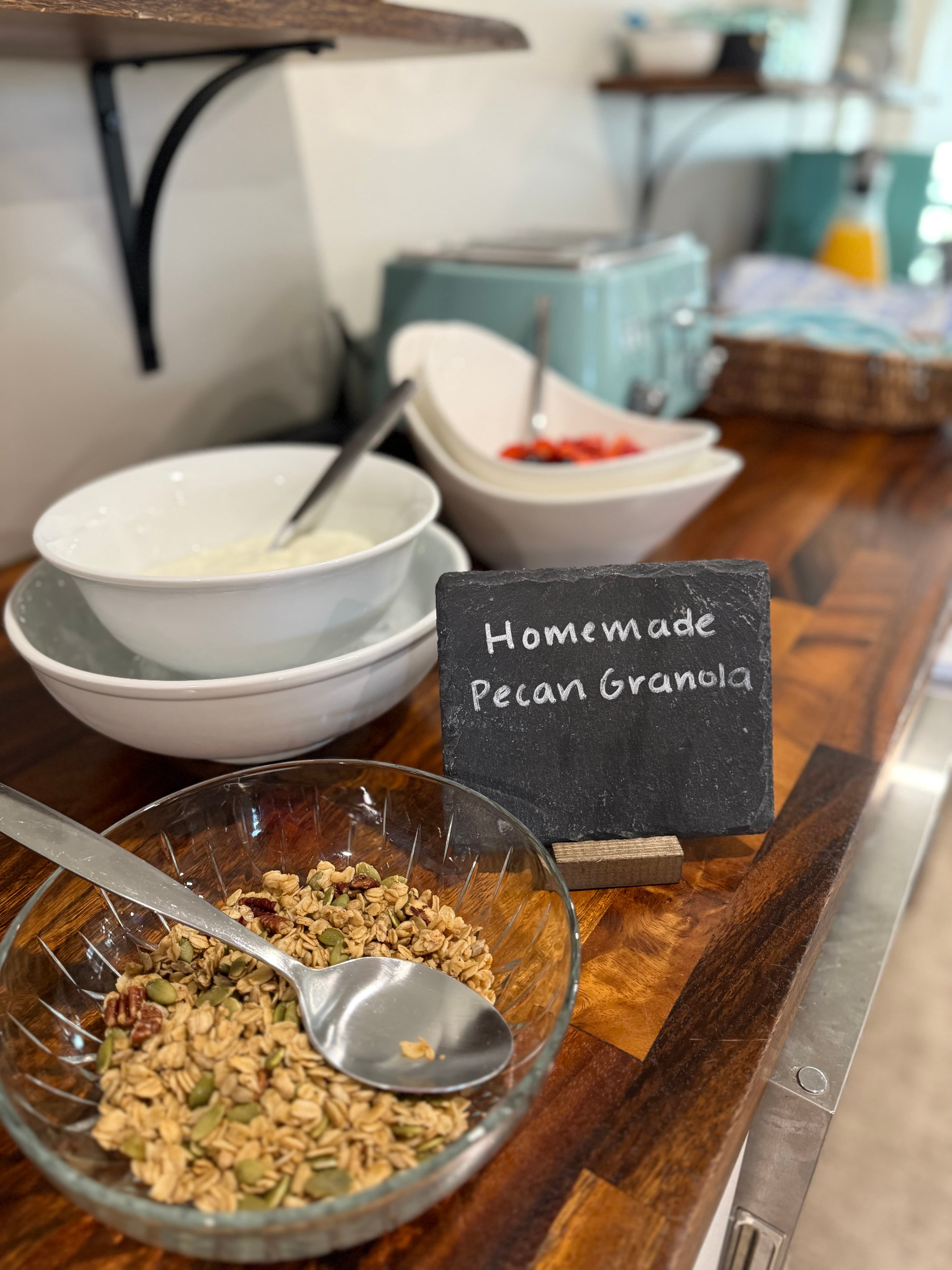 A bowl of homemade pecan granola with a spoon, accompanied by other serving dishes on a wooden counter.