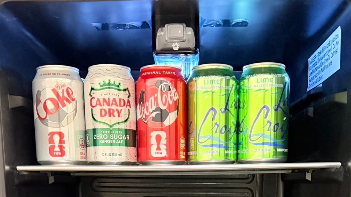 A variety of soft drink cans, including Coke, Canada Dry, and La Croix, are displayed in a fridge.