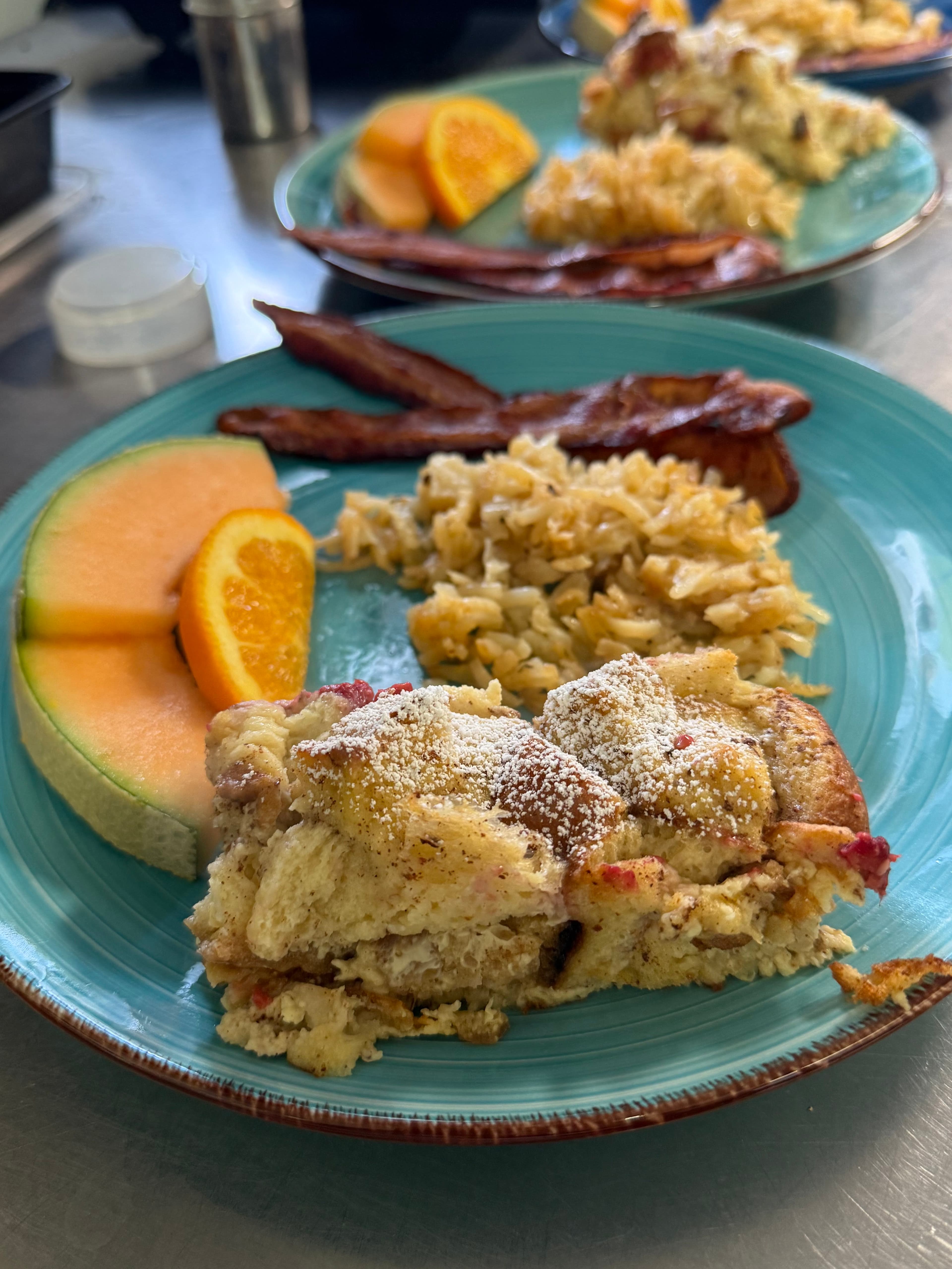 A blue plate with baked French toast, scrambled eggs, bacon, and melons.