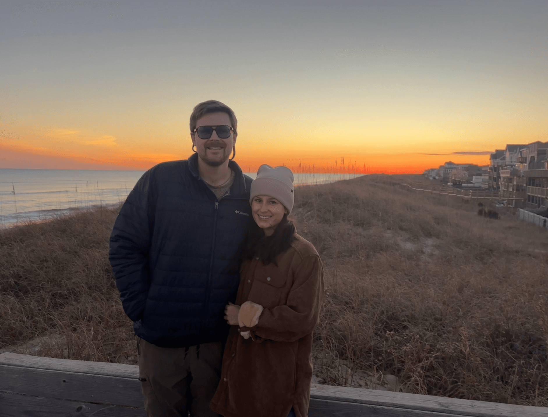A couple stands together on a beach at sunset, with soft waves and grassy dunes in the background.