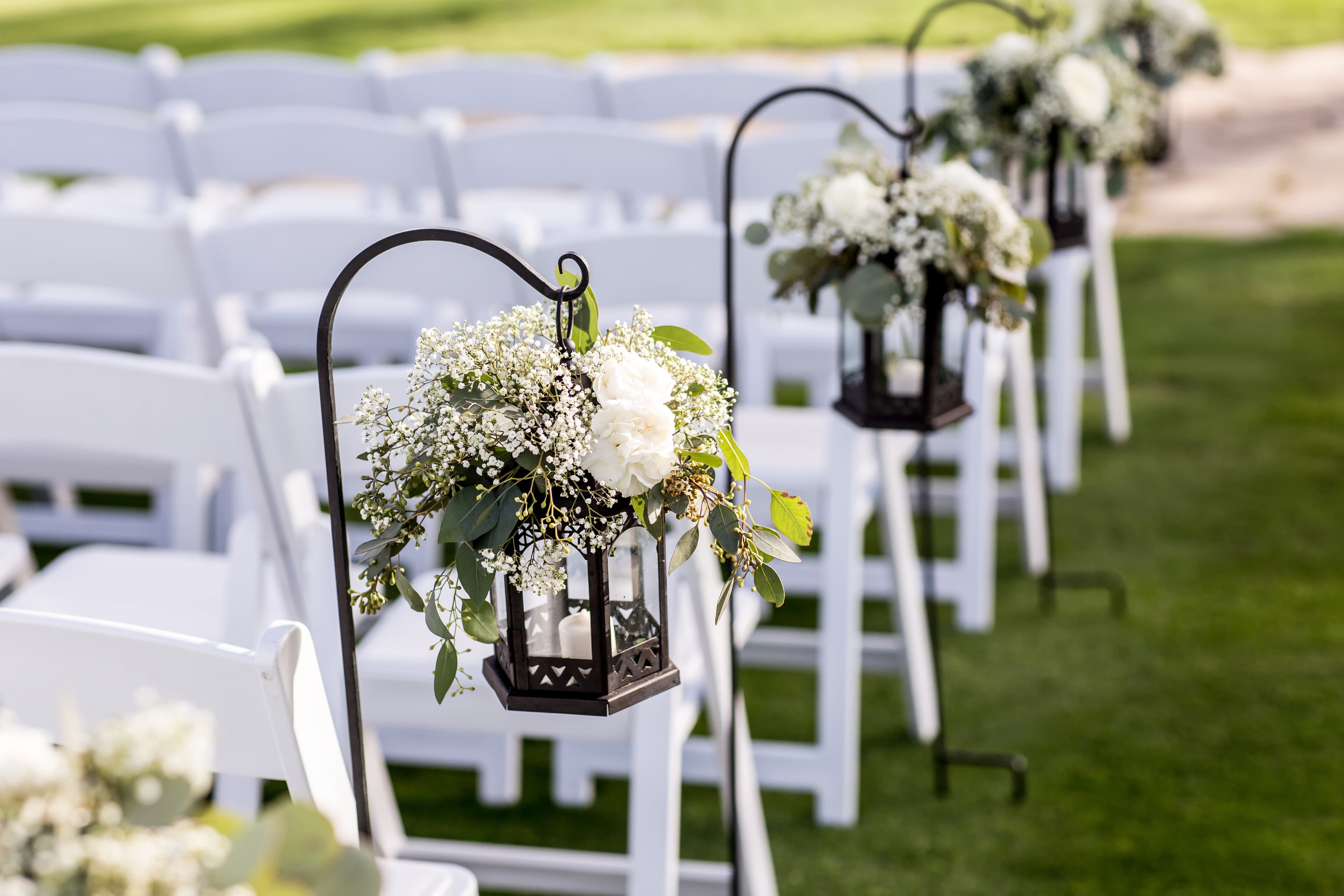 Black lanterns with floral arrangements hang from white chairs at an outdoor wedding venue.