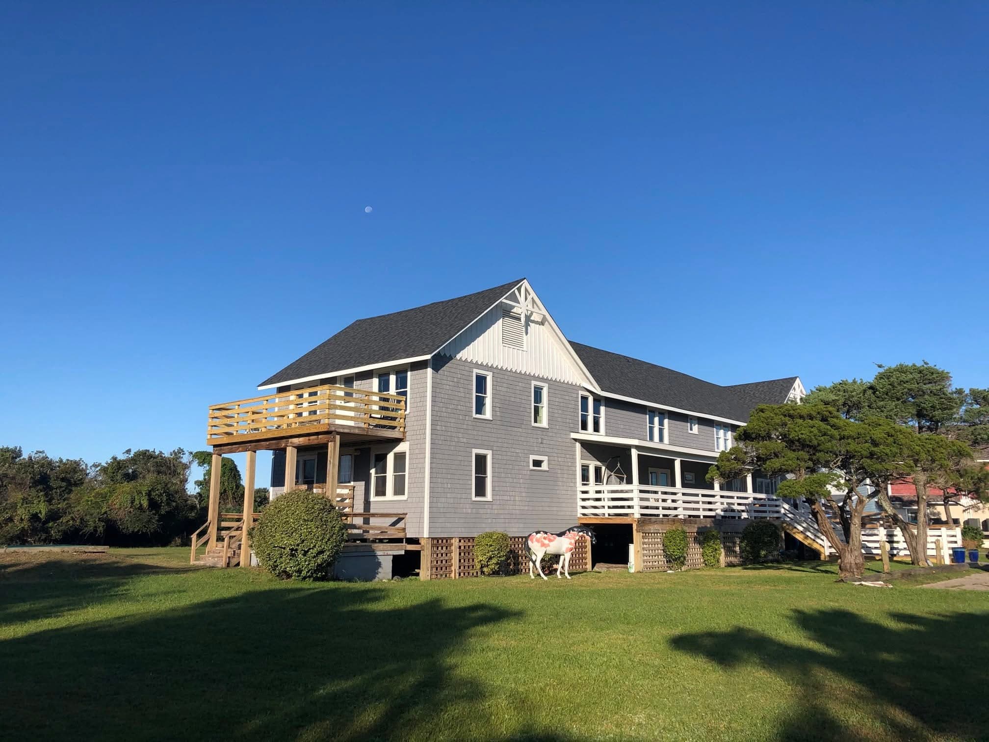 A large gray house with a wooden deck, set on green grass under a clear blue sky.