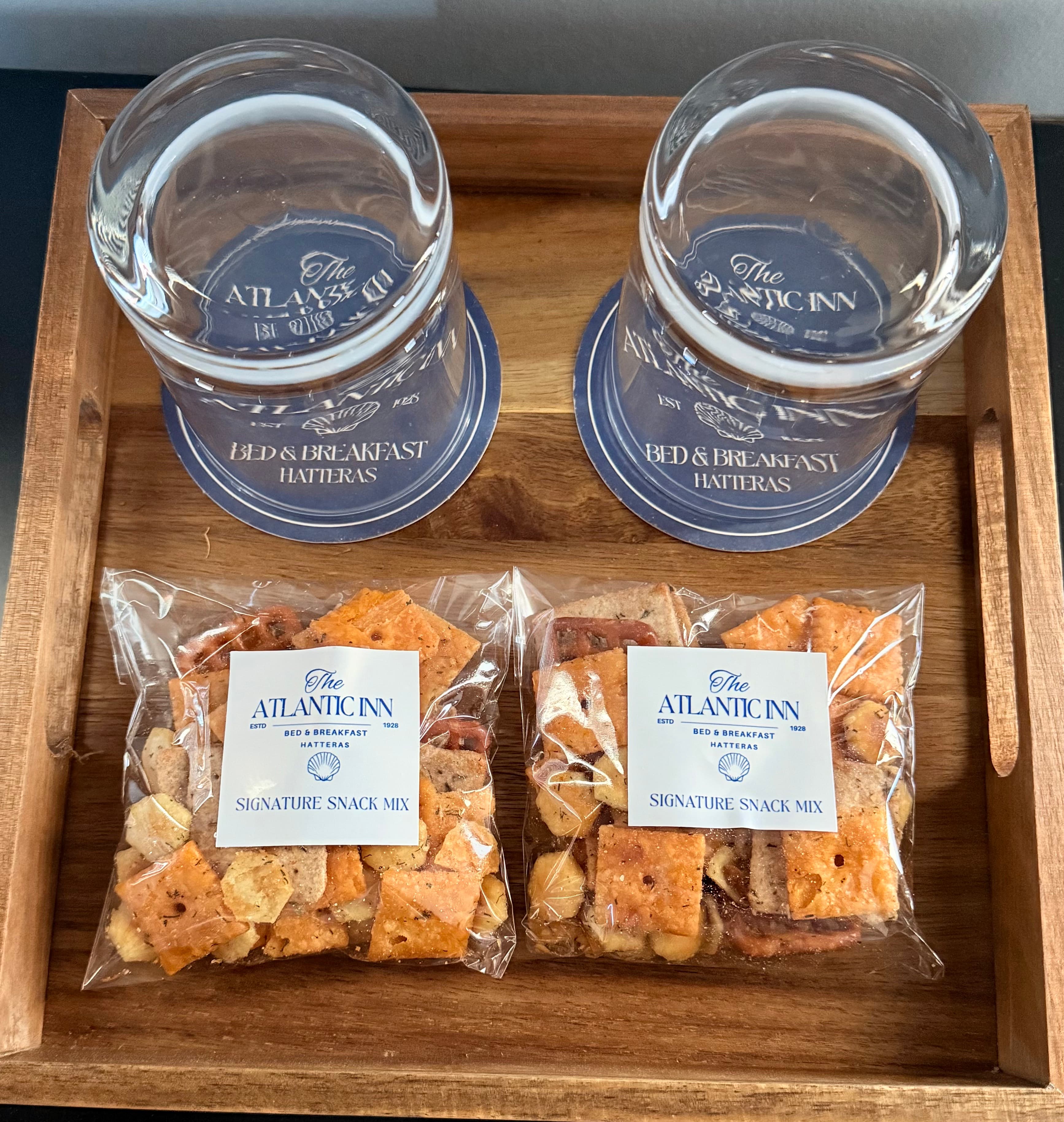 A wooden tray holds two glasses, coasters, and two bags of signature snack mix from The Atlantic Inn.