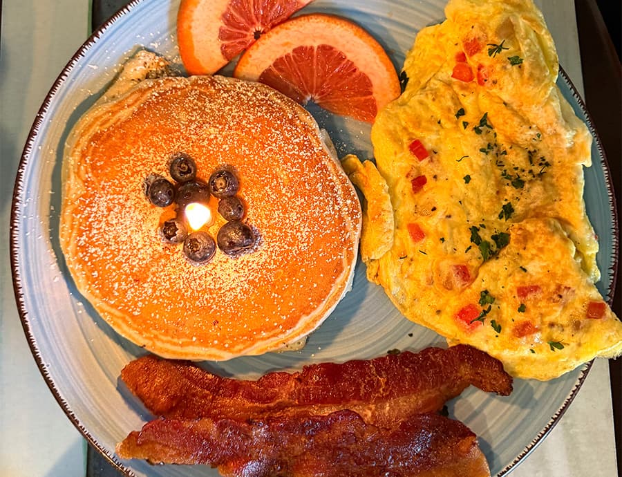 A plate of breakfast featuring a stack of pancakes with blueberries and powdered sugar, an omelet with diced vegetables, crispy bacon, and slices of grapefruit.