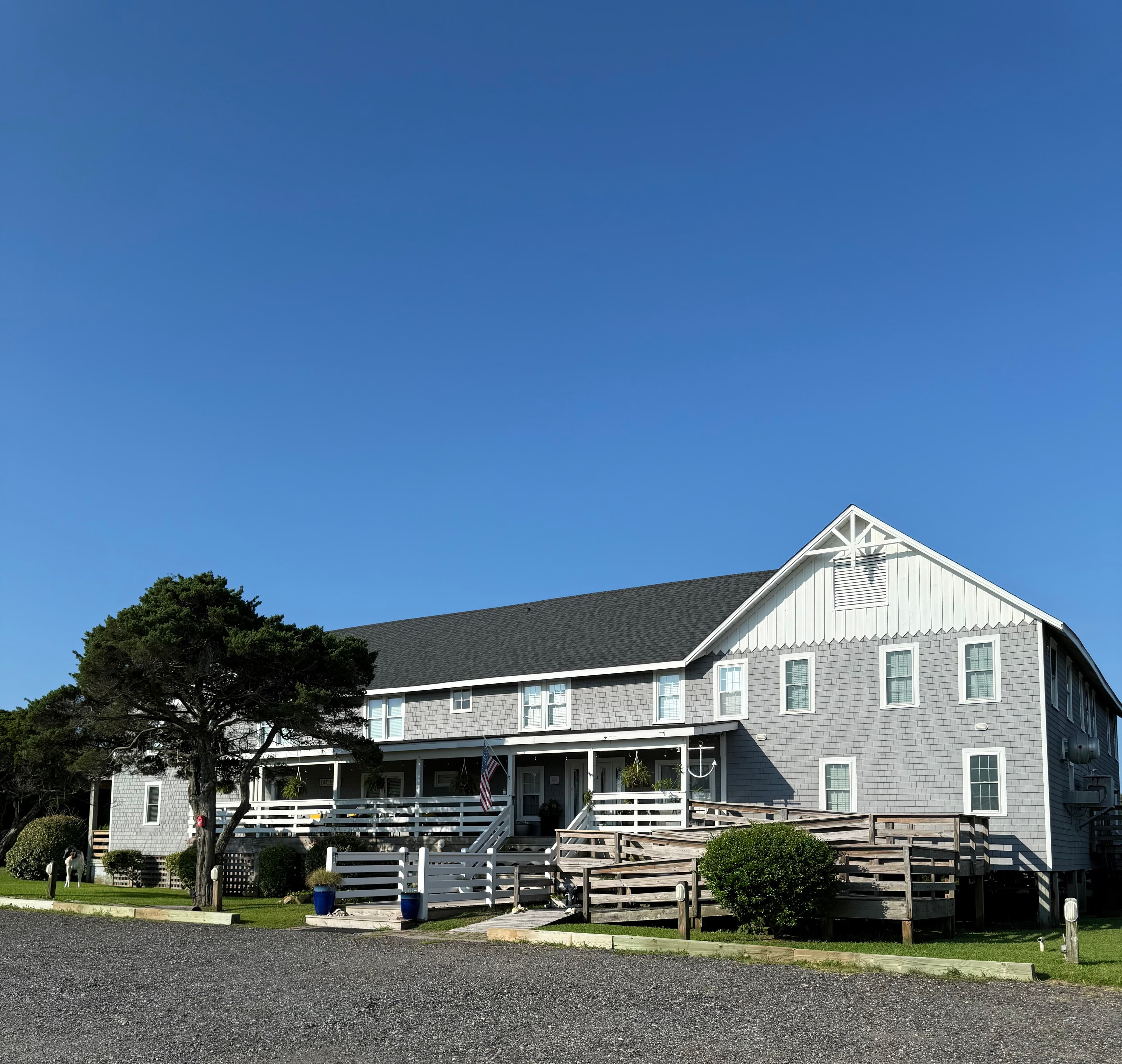 A gray building with a gabled roof and front porch, surrounded by greenery under a clear blue sky.