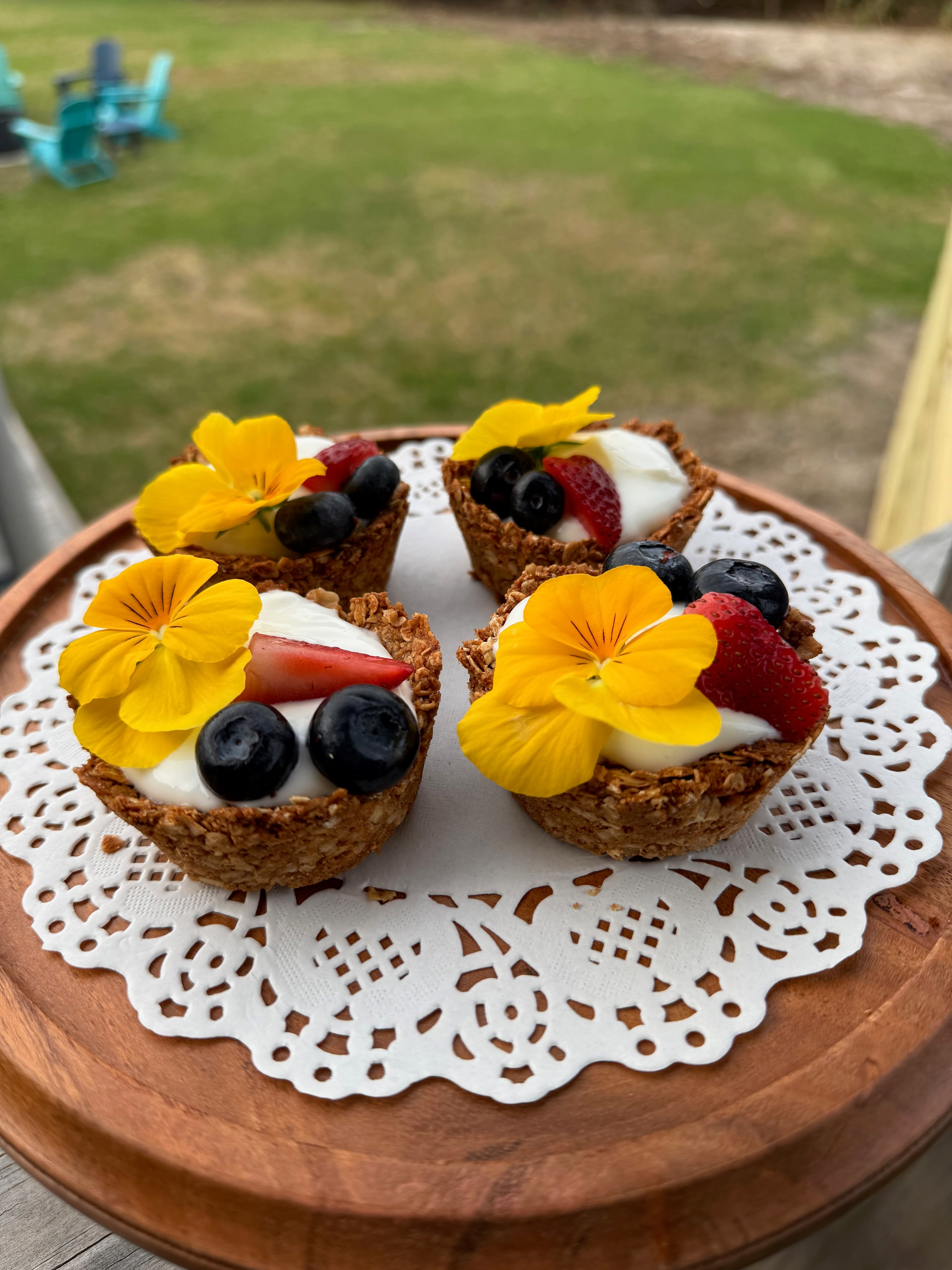 Four mini fruit tarts topped with cream, fresh berries, and edible flowers, presented on a lace doily.