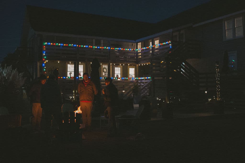 People gather around a small fire outside a brightly lit building at night.