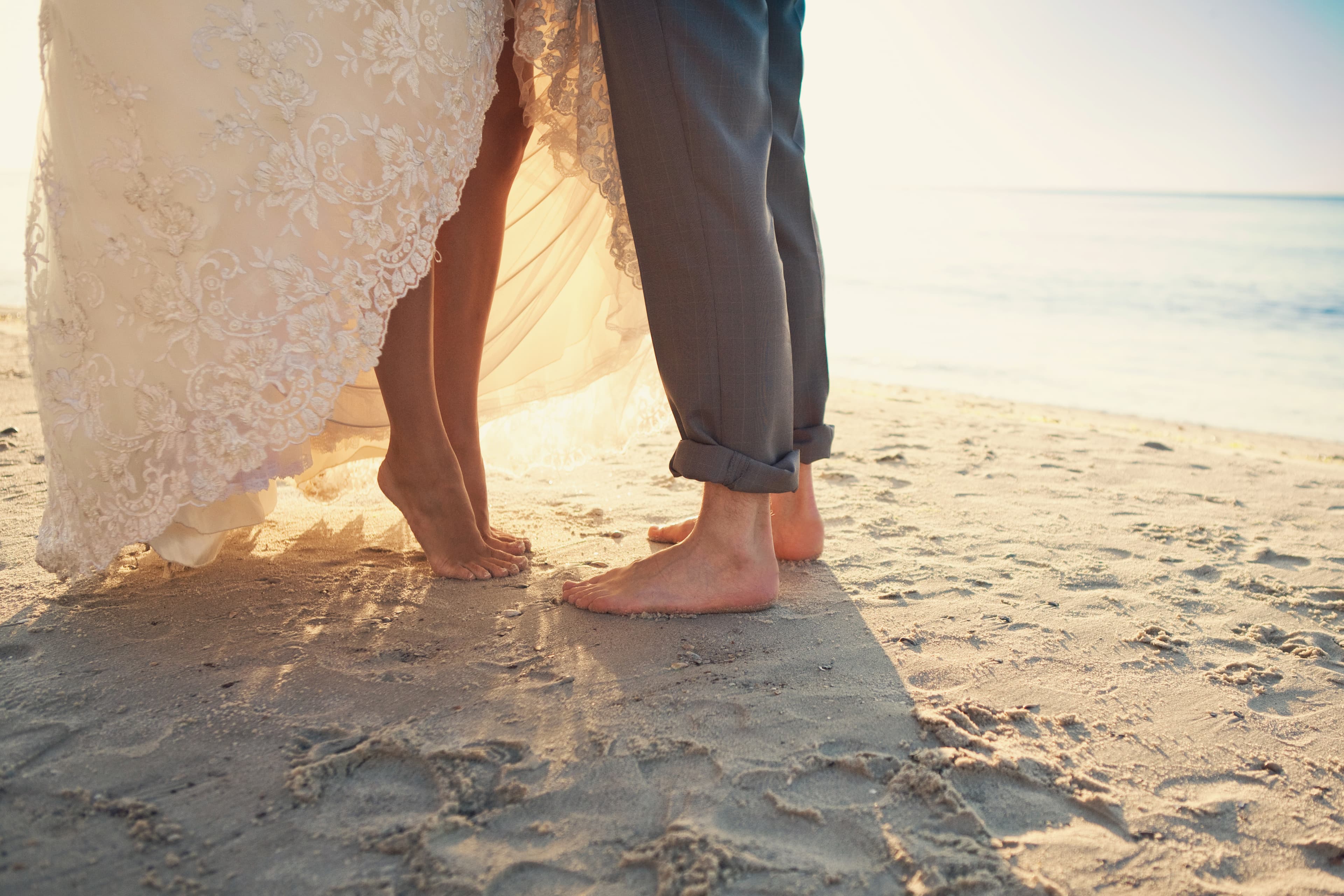 A couple stands barefoot on the beach, their feet close together in the soft sand.