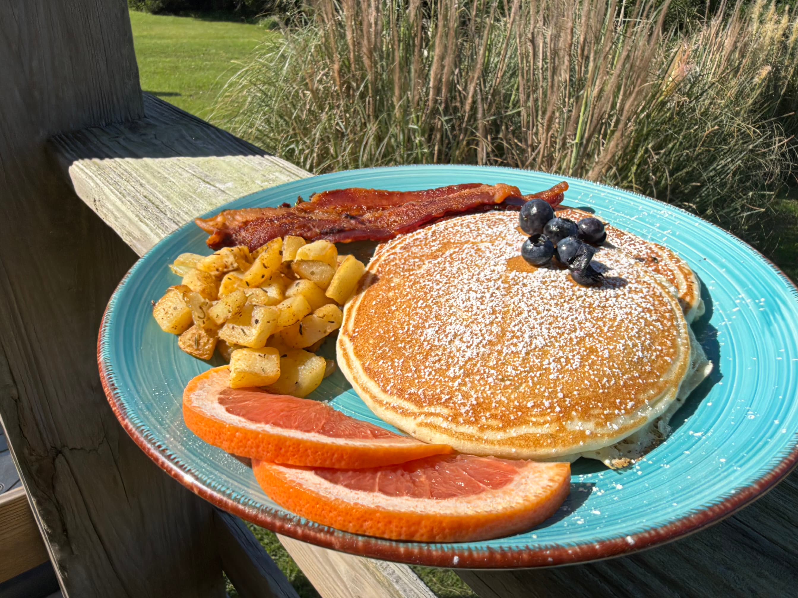 A plate of pancakes topped with blueberries, served with bacon, seasoned potatoes, and grapefruit slices, set outdoors.