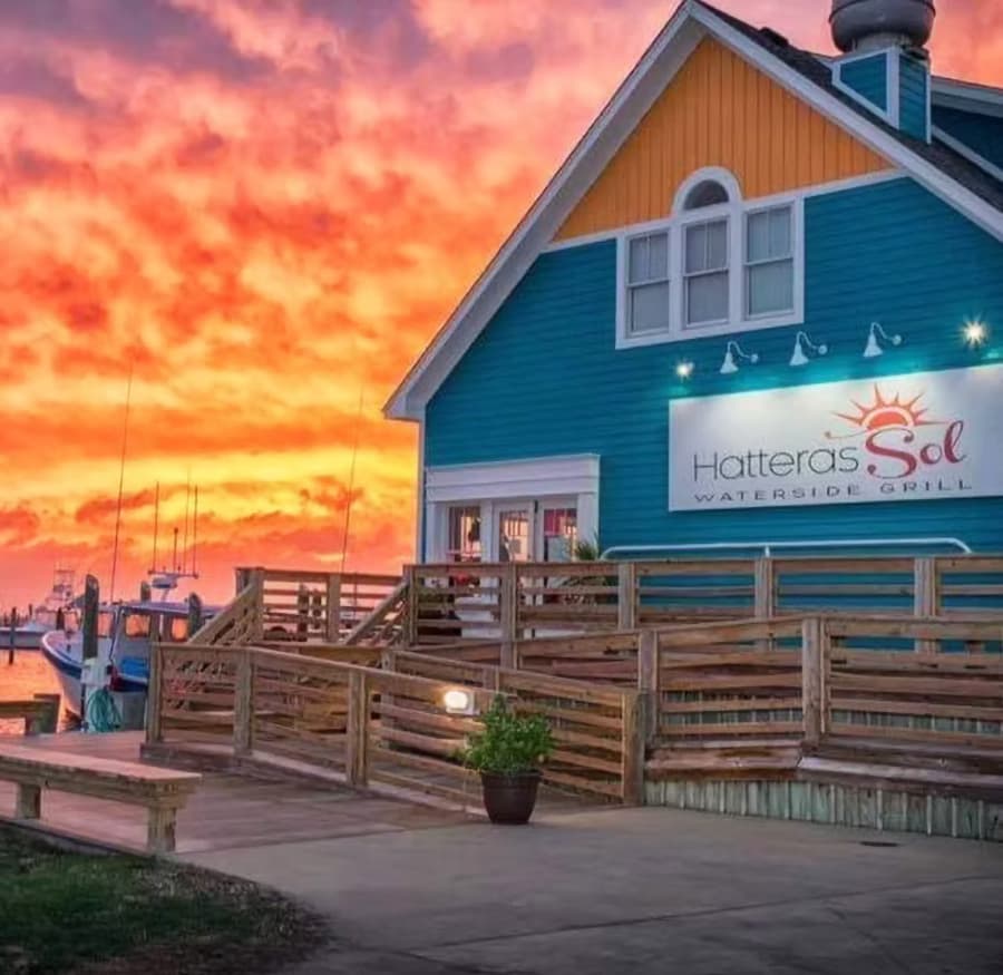 Colorful sunset over the Hatteras Sol Waterside Grill, a blue building with wooden walkways.