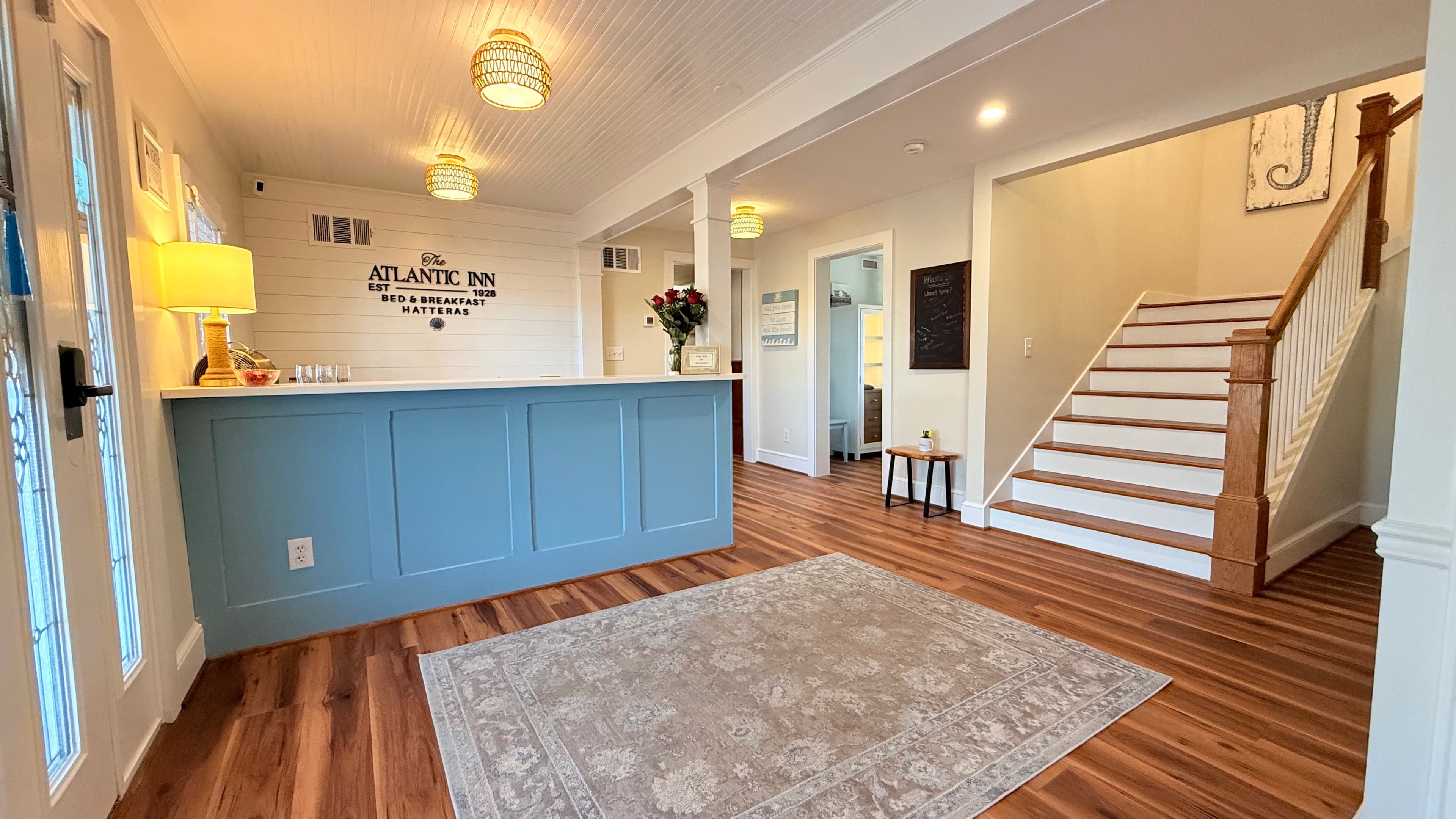 Interior view of a warmly lit reception area with a blue front desk, wooden flooring, and a staircase in the background.