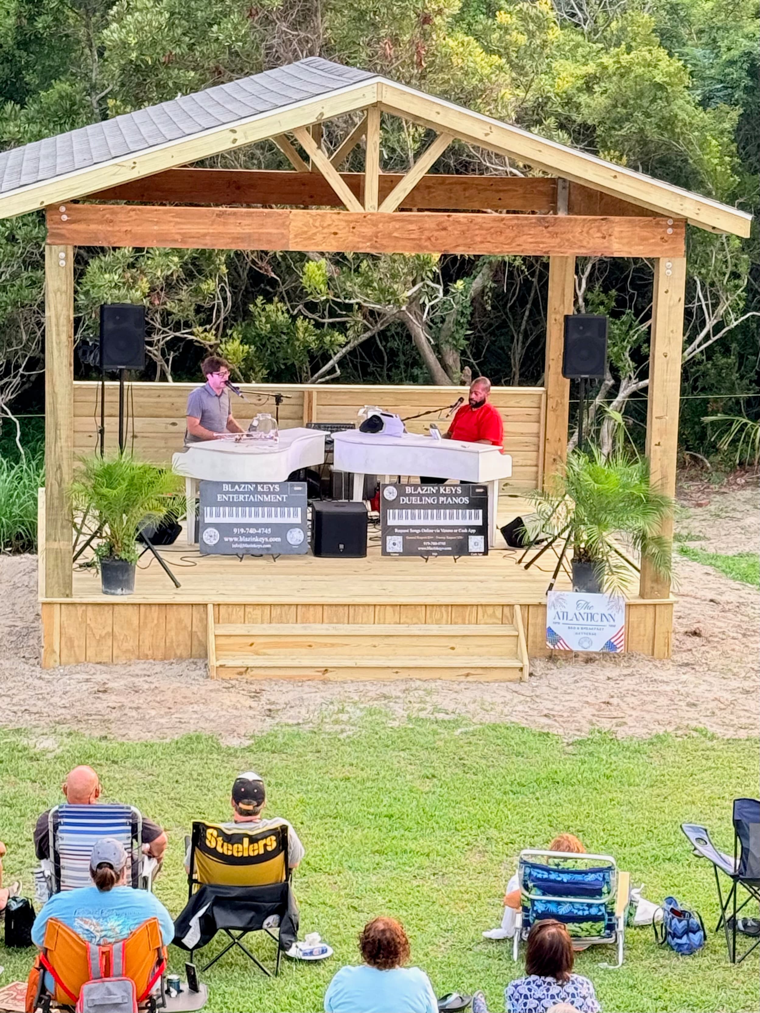 Two performers play dueling pianos on a wooden stage while an audience watches from lawn chairs.