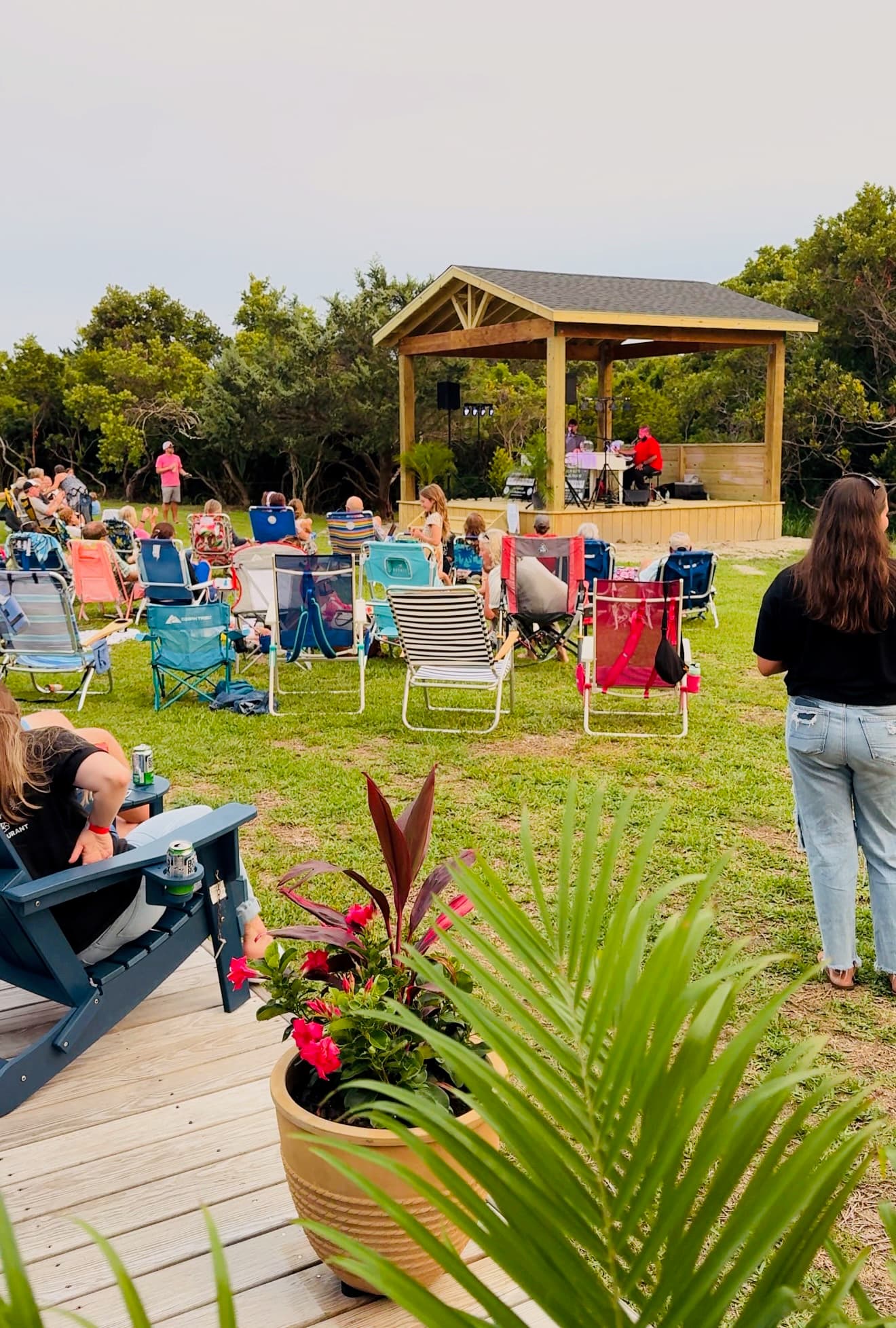 Audience members seated in portable chairs enjoy a live performance at an outdoor stage surrounded by greenery.