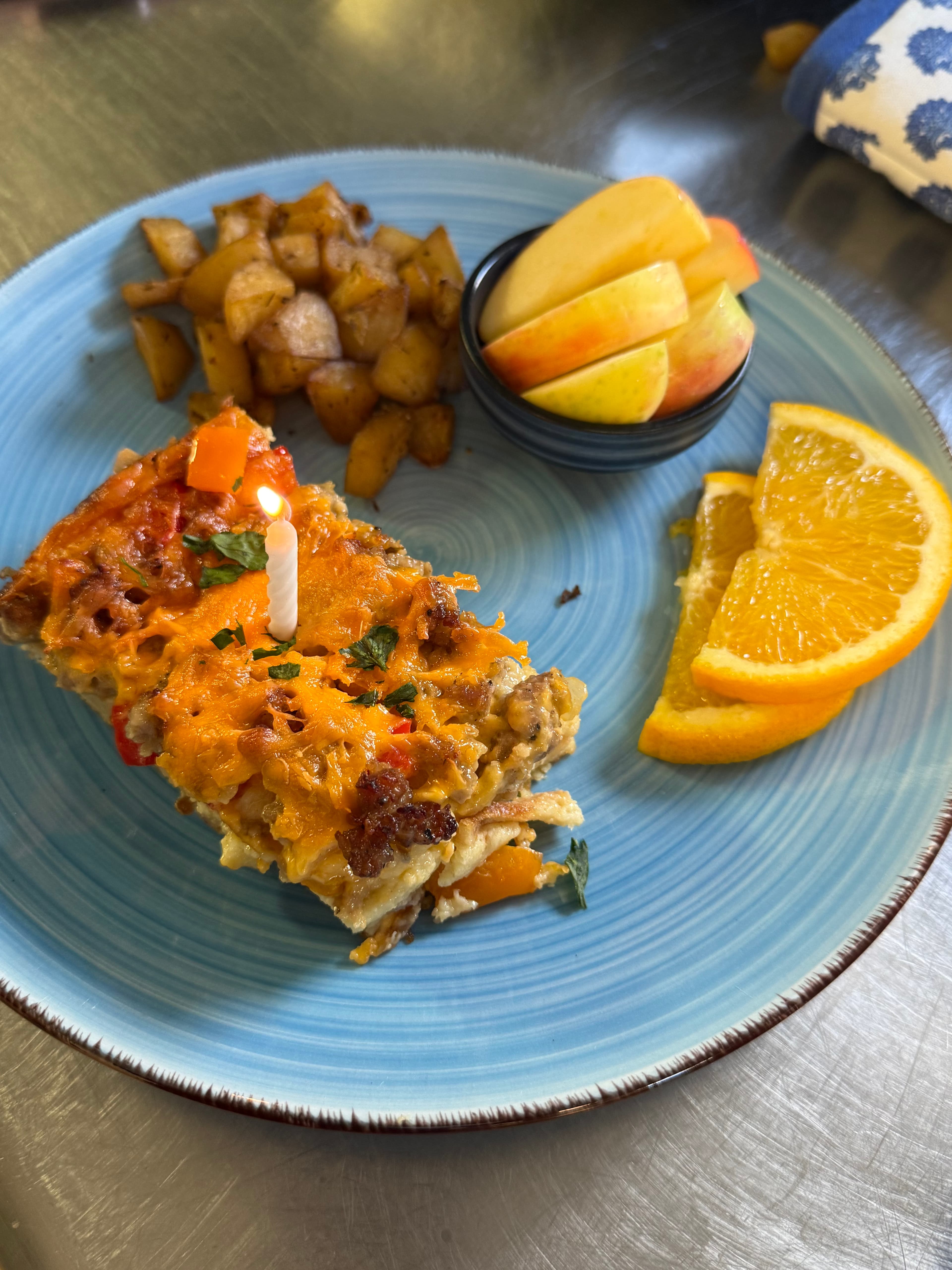 A colorful breakfast dish with a slice of cheesy casserole, a birthday candle, seasoned potatoes, apple slices, and orange wedges on a blue plate.