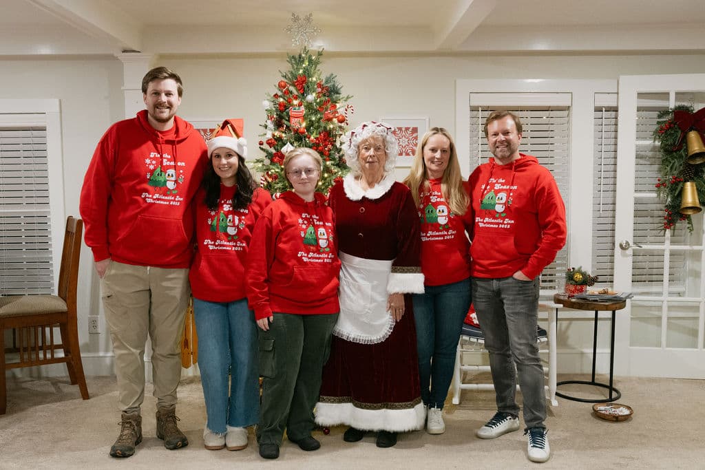 A group of six people in festive attire poses together in a cozy room decorated for Christmas.